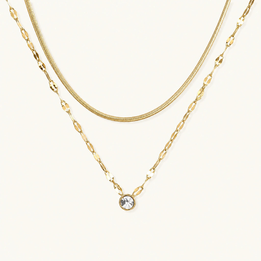 Layered gold necklace with herringbone and link chains, small bezel-set crystal pendant on cream background