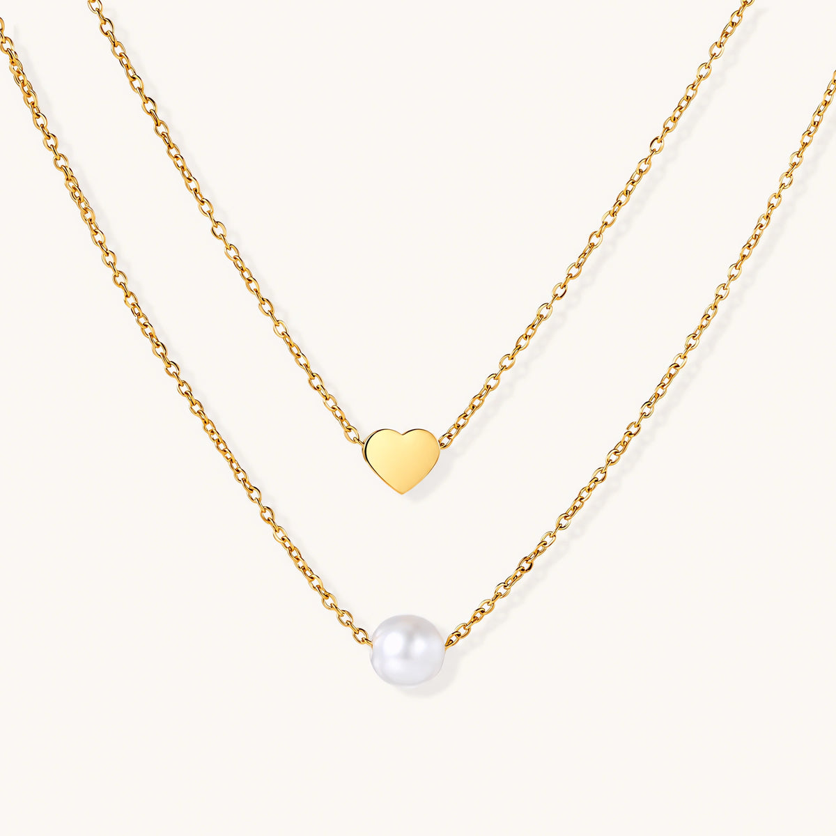 Two delicate gold-plated chains with heart charm and white pearl bead on cream background, minimal layered design.