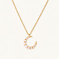 Gold crescent moon pendant with graduated white pearls on delicate chain against cream background