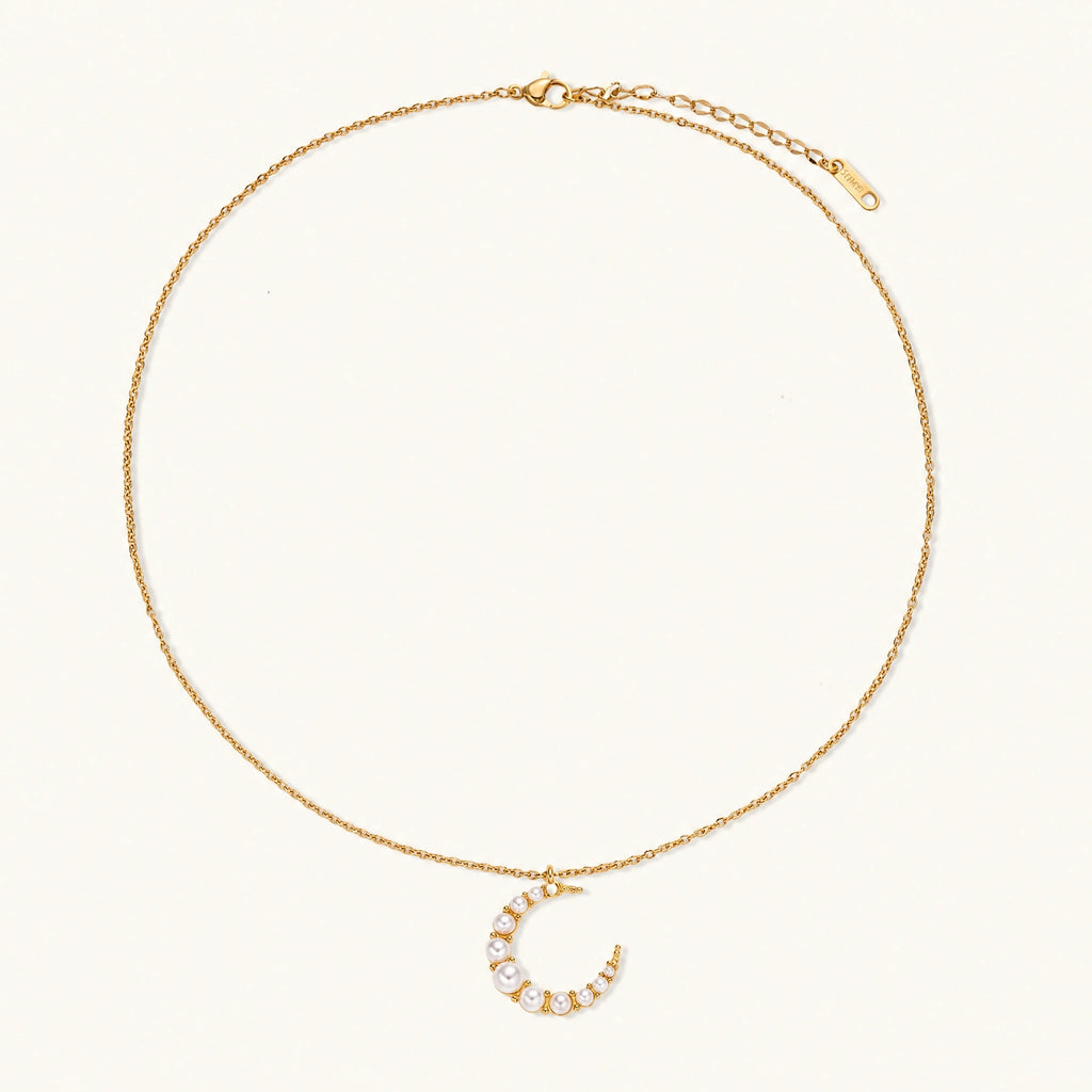 Gold crescent moon pendant with graduated white pearls on delicate chain against cream background