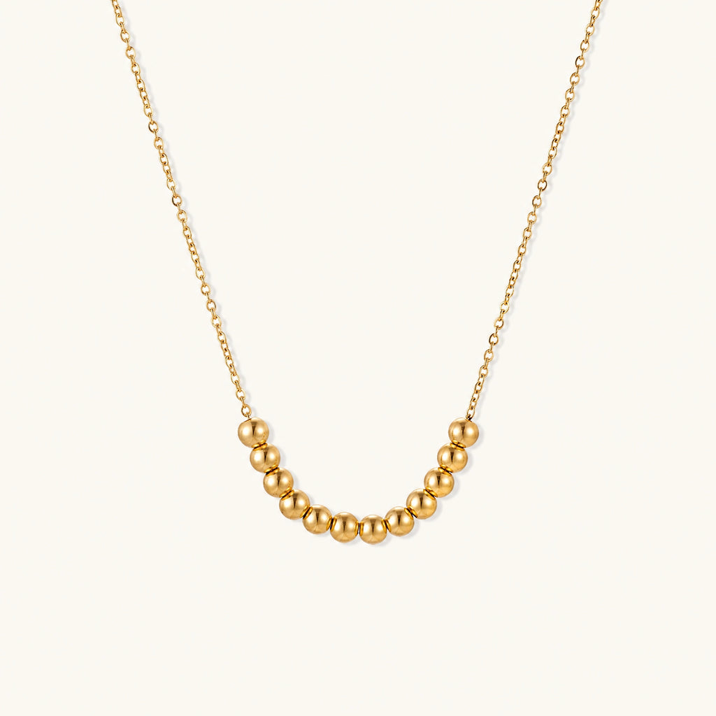 Delicate gold-plated necklace with graduated spherical beads forming gentle smile curve on beige background