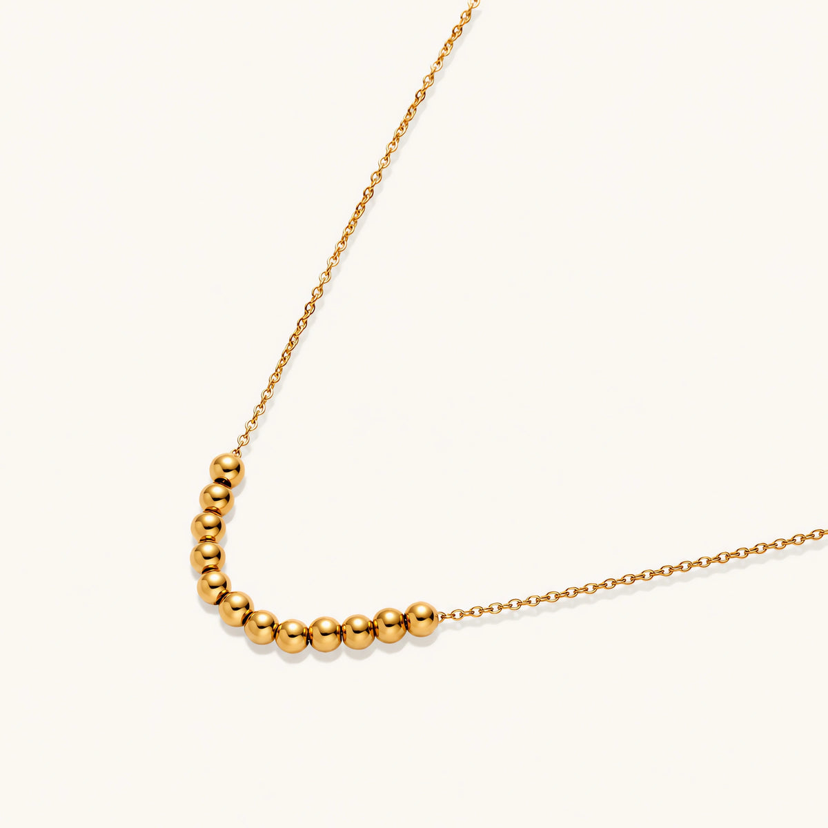 Delicate gold-plated necklace with graduated spherical beads forming gentle smile curve on beige background