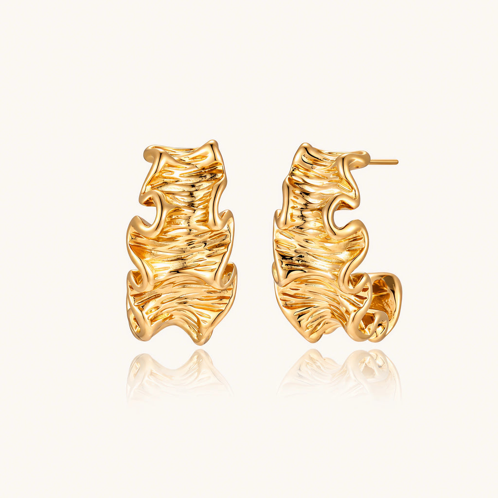 Pair of gold huggie hoops with sculpted wave texture and dimensional ripples on neutral background.