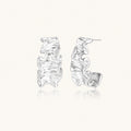 Textured white gold-plated hoop earrings with flowing wave pattern against cream background, showing dimensional rippled surface.