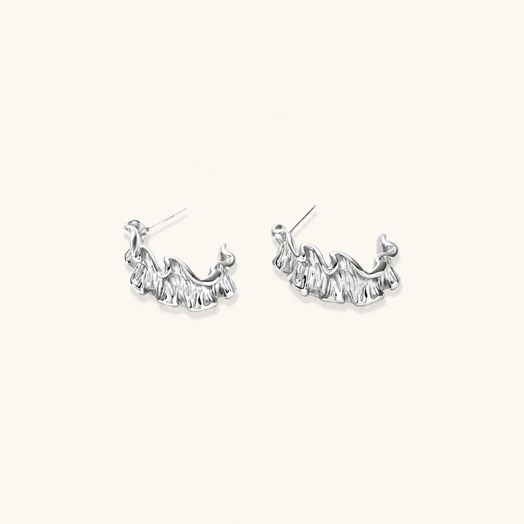 Textured white gold-plated hoop earrings with flowing wave pattern against cream background, showing dimensional rippled surface.