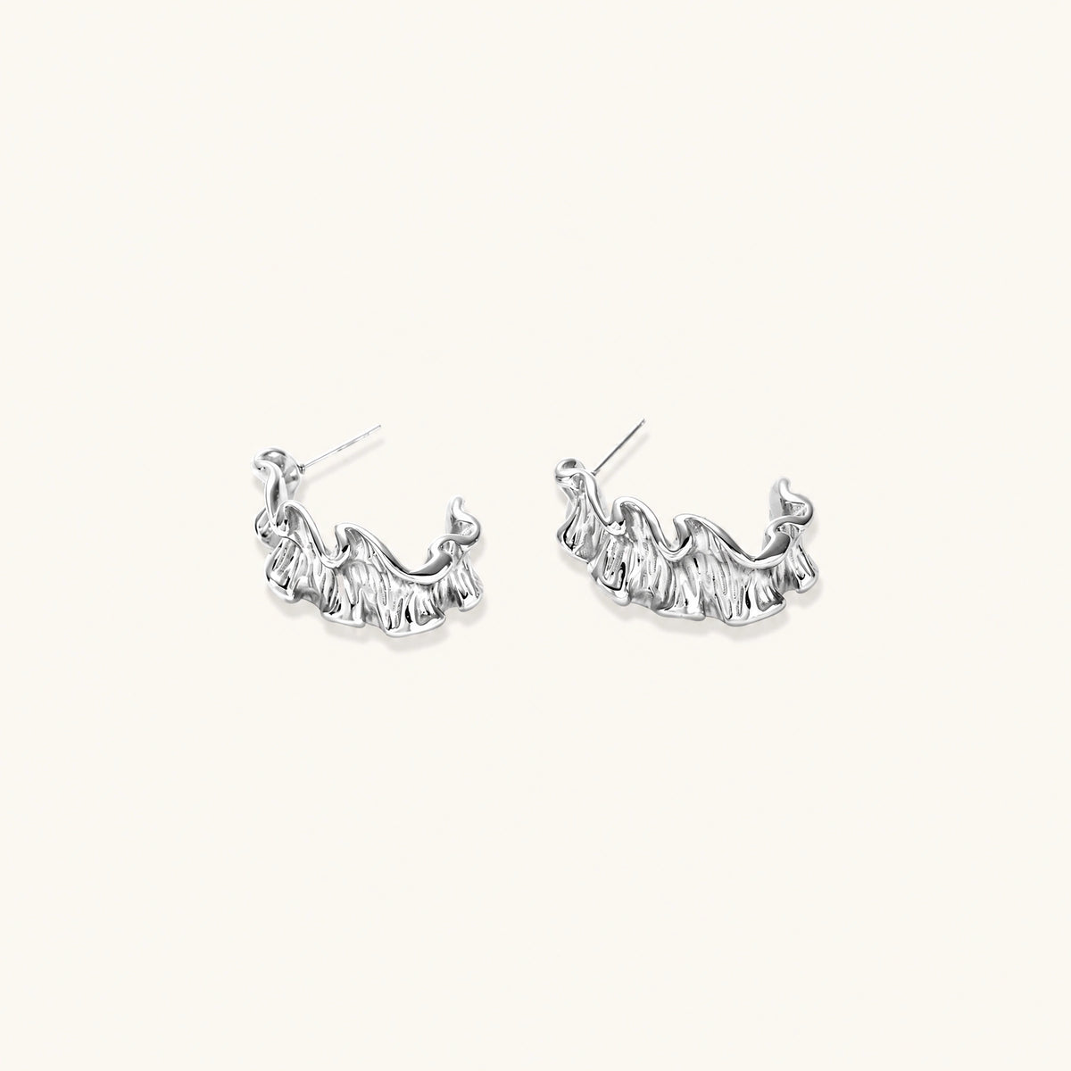 Textured white gold-plated hoop earrings with flowing wave pattern against cream background, showing dimensional rippled surface.