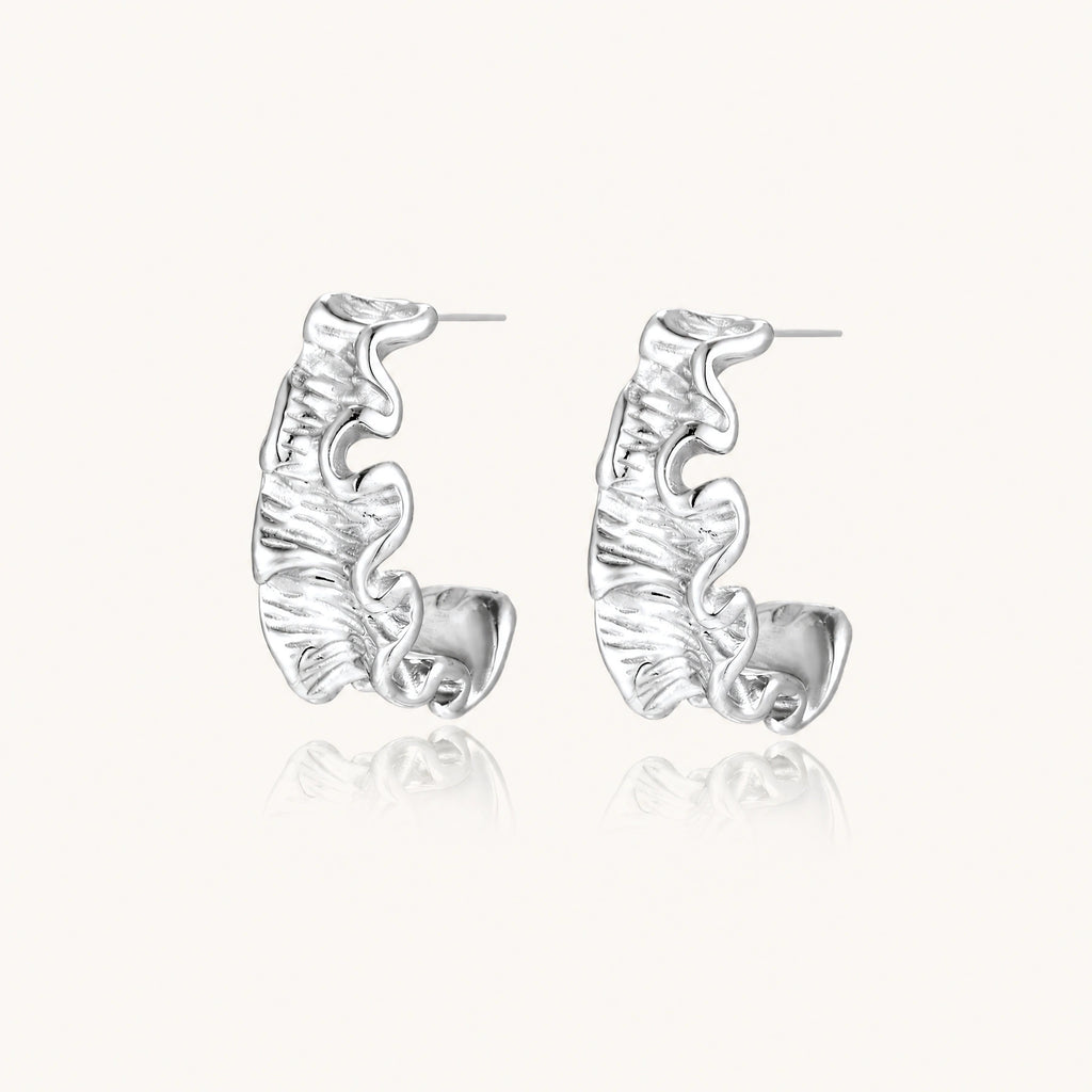 Textured white gold-plated hoop earrings with flowing wave pattern against cream background, showing dimensional rippled surface.