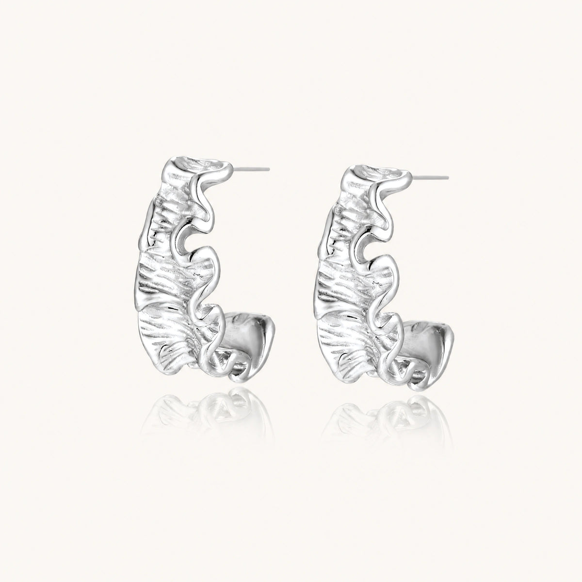 Textured white gold-plated hoop earrings with flowing wave pattern against cream background, showing dimensional rippled surface.