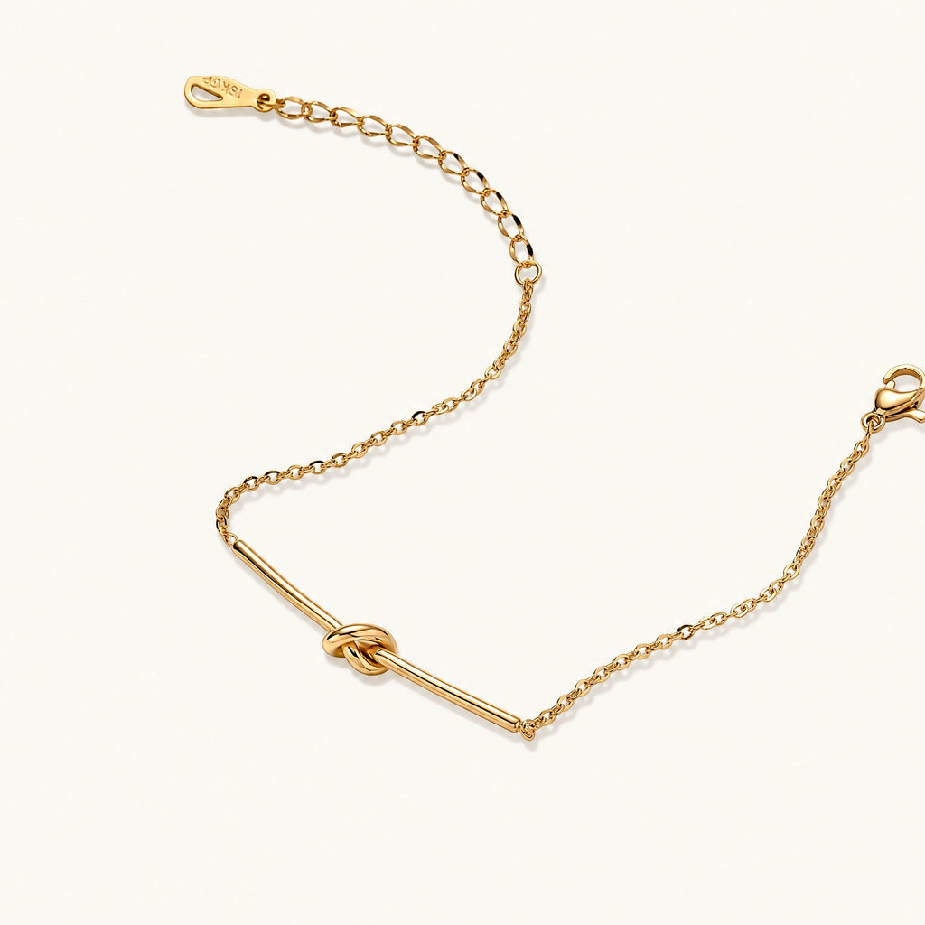 Delicate gold chain bracelet with minimalist bar and centered knot detail on cream background