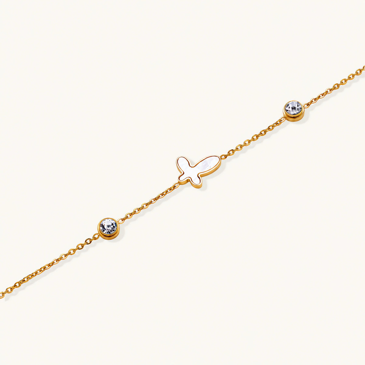 Gold chain bracelet with white mother of pearl butterfly pendant and two bezel-set crystals on cream background