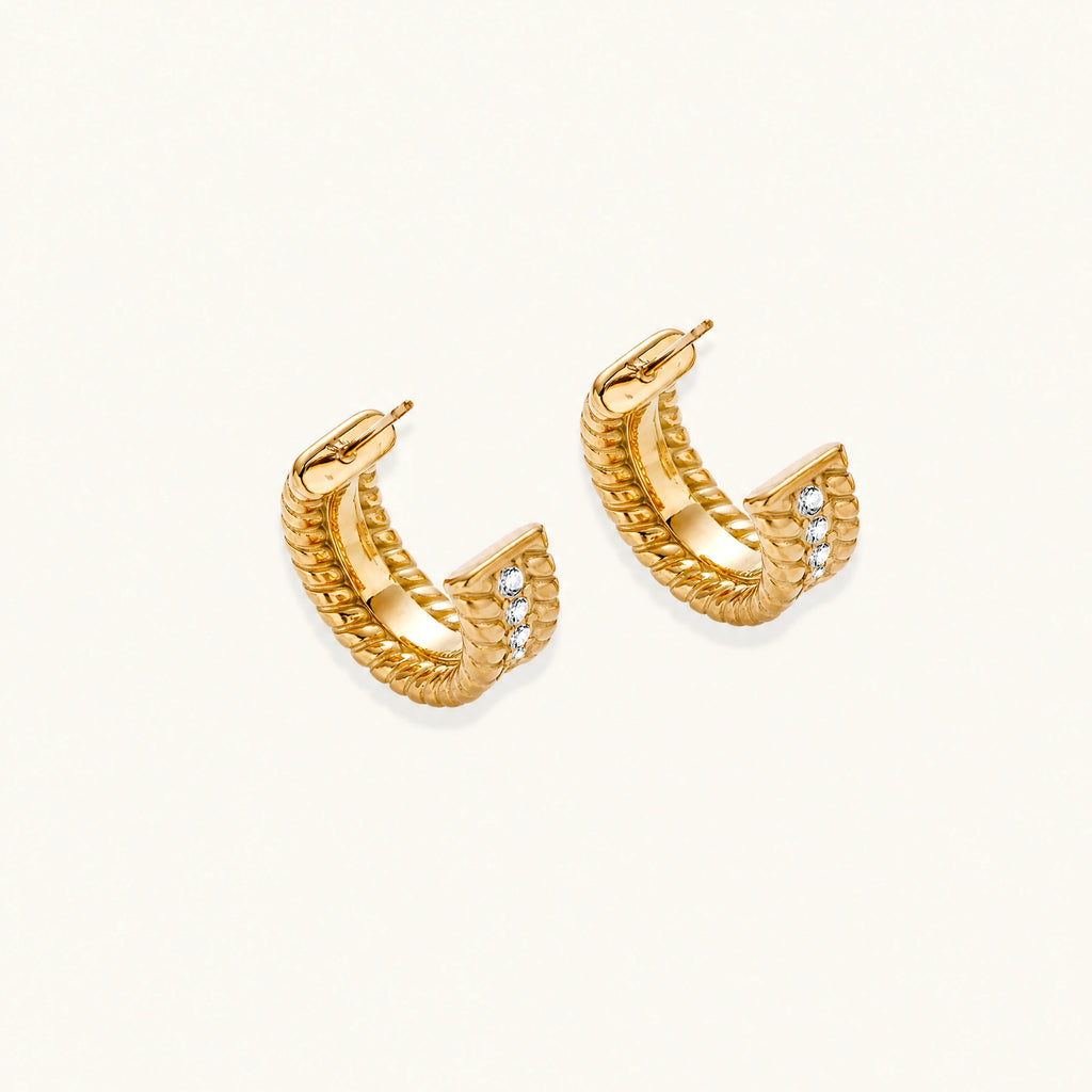 Textured gold huggie hoop earrings with raised ribbed rows creating dimensional sculptural design on cream background